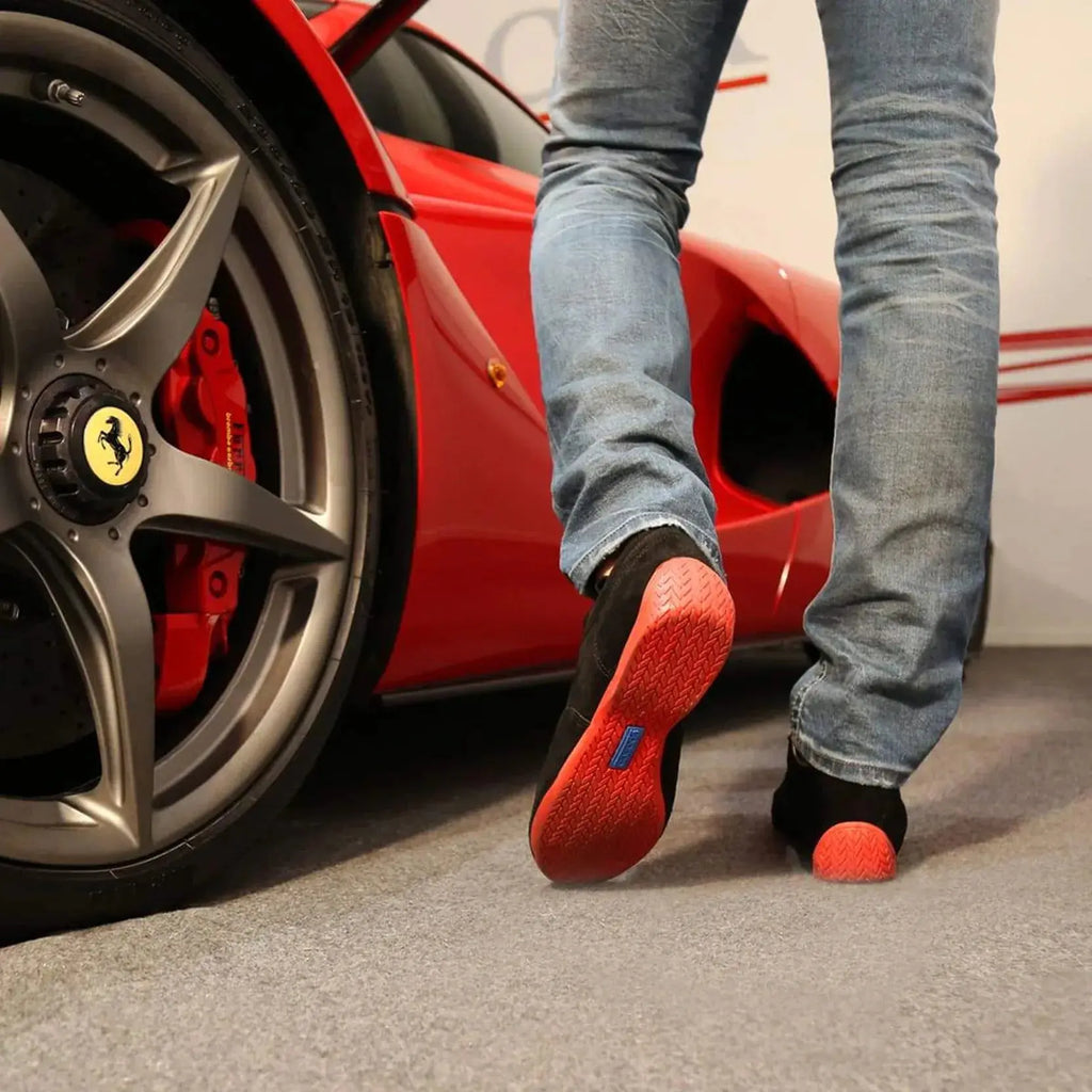 Paire de chaussures Sparco Imola en cuir suédé noir avec semelle rouge, portée de dos, devant une Ferrari