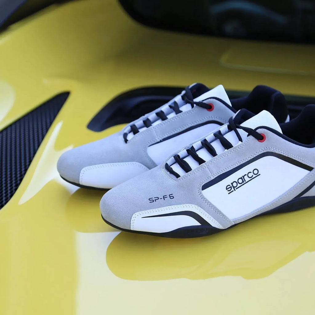 Paire de Sneakers Sparco SP-F6 blanc et gris, lacets noirs, cuir et suède, semelle noire qui remonte sur le talon, Sparco sur le côté, posée sur un capot